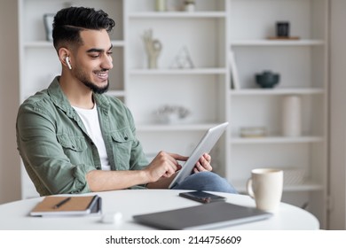 Modern Technologies. Smiling Young Arab Guy Using Digital Tablet At Home, Happy Millennial Middle Eastern Man In Airpods Sitting At Desk And Browsing Internet On Modern Gadget, Copy Space - Shutterstock ID 2144756609