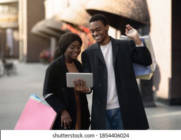 Modern technology, cashback and sale message. Cheerful millennial african american man in coats holds shopping bags and shows digital tablet to his wife on street near boutique, outdoor, copy space