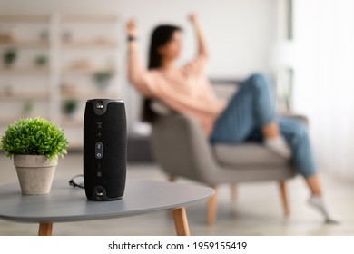 Modern Technology Concept. Closeup of smart portable wireless speaker on the table, selective focus. Young woman listening to music and dancing, sitting on armchair in the blurred background - Shutterstock ID 1959155419