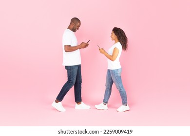 Modern technology and relationship. Full body length of black middle aged man and young woman using their smartphones, texting each other, walking over pink studio background, banner