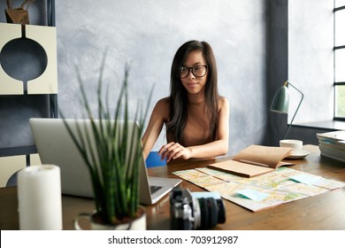 Modern technology, travel, tourism and vacations concept. Attractive positive young Chinese woman travel agent sitting at her office desk in front of laptop, booking journey; copybook and map on table