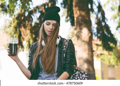 Modern teenage hipster girl in autumn in park in beanie hat, with takeaway coffee and headphones Millennial young woman in fall relaxing, drinking cappuccino. Matte filter, natural lighting.
