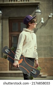 A Modern Teenager Boy In A Hoodie And Sunglasses Is Standing On The Street With A Longboard In His Hand. Activity, Summer And Youth Lifestyle.