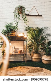 Modern Terrace Decorations With Indoor And Outdoor Plants And Flowers, Wooden Table, Clay Pots, Seagrass Baskets And Rug With Led Lights On A White Brick Wall During Early Summer Evening