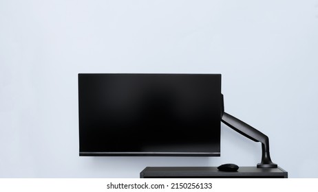 Modern thin computer monitor or TV set is mounted on a metal swivel desktop bracket with a gas lift function. Small black table. Off-center side holder. Copyspace. Light background