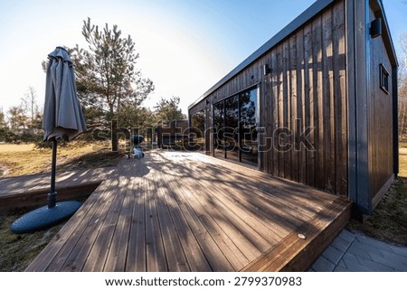 Modern timber cabin with expansive deck and umbrella stand in late afternoon light. Wood clad structure with sliding glass doors sits by young pines and grill area.