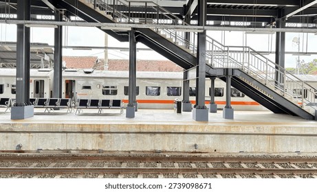 A modern train station platform features a stationary train, empty waiting benches, and a prominent metal staircase leading to an upper level, with railway tracks visible in the foreground. 