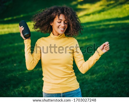 Modern trendy african american girl listening to music by wireless portable speaker.Young beautiful woman enjoying, dancing in park.