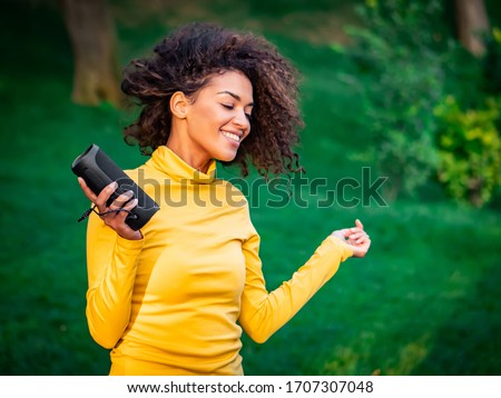 Modern trendy african american girl listening to music by wireless portable speaker.Young beautiful woman enjoying, dancing in park.