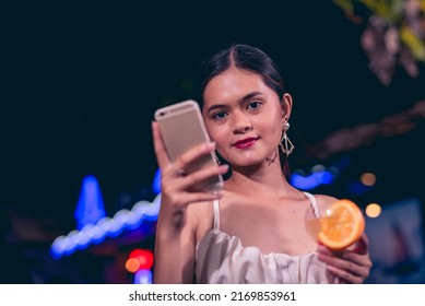 A modern and trendy young woman checking her social media while drinking a glass of tequila sunrise. Vibrant nightlife outdoor scene at a bar or cafe with neon lights. - Powered by Shutterstock