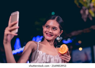 A modern and trendy young woman taking a selfie of herself with a glass of tequila sunrise. Vibrant nightlife outdoor scene at a bar or cafe with neon lights. - Shutterstock ID 2169853953