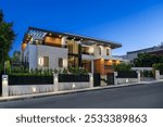 Modern two-story house with a sleek design, illuminated exterior, and landscaped front yard during twilight.