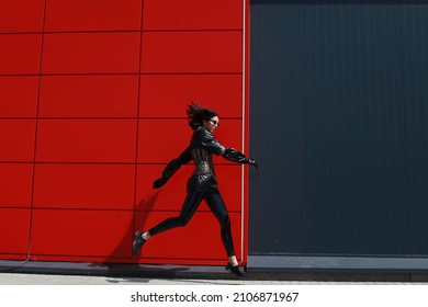 Modern urban lifestyle. Stylish young woman in trendy tight minimalist latex outfit and sunglasses is jumping on run on a street by the red industrial wall. Female fashion. Copy space for ad.

