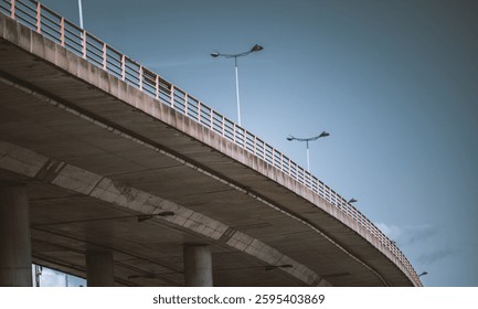 Un moderno paso elevado urbano con pilares de hormigón y barandillas metálicas, capturado desde una perspectiva de ángulo bajo. Farolas con brazos curvos alinean el puente contra un cielo azul claro
