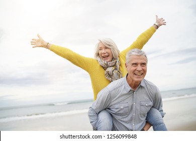 Modern vibrant senior couple piggy back riding on beach                               