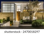 A modern villa with a stone facade and large windows is illuminated at night. A large olive tree stands in front of the entrance. The walkway leading  is lined with green shrubs and bushes.