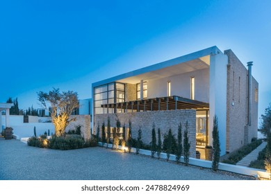 Modern Villa With Stone Facade and Lush Greenery at Dusk. The villa has a large patio with a pergola, and the windows are all lit up. The landscaping is well-maintained, with a variety of trees - Powered by Shutterstock