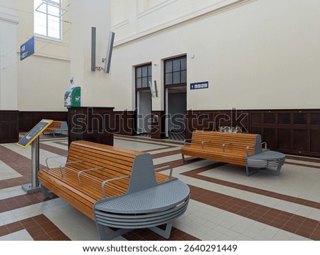 Modern waiting area inside a train station with wooden benches, power outlets, a defibrillator, and directional signs. The clean and bright hall features classical architecture and tiled flooring.