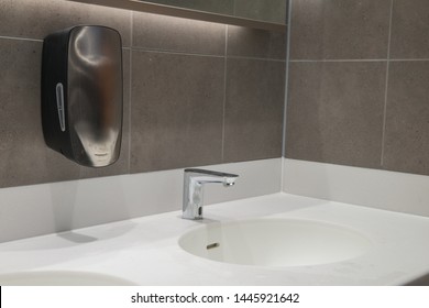 Modern Wash Hand Basin With Hand Free Sensor Tap And Soap Dispenser. Solid Surface Worktop And Grey Tiled Wall.