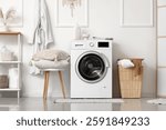 Modern washing machine with laundry basket and pillows on stool near white wall in bathroom