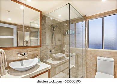 Modern Washroom, Shower Area And Washing Area, Including A White Sink And Silver Taps On The Ceramic Counter Beside Glass Cover And The Toilet Bowl, The Mirror Is Attached To The Wall