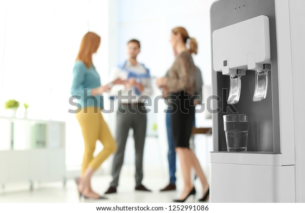 Modern water cooler with glass and blurred\
office employees on background. Space for\
text