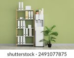 Modern water cooler with paper cups and shelf unit near green wall in office