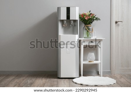 Modern water cooler and vase with flowers on end table near grey wall