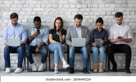 Modern way to communicate. Row of diverse young people sitting close to wall holding different gadgets cellphones laptops pads surfing internet chatting browsing social networks work or study online