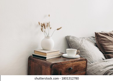 Modern white ceramic vase with dry Lagurus ovatus grass and cup of coffee on retro wooden bedside table. Beige linen and velvet pillows in bedroom. Scandinavian interior. Homestaging.