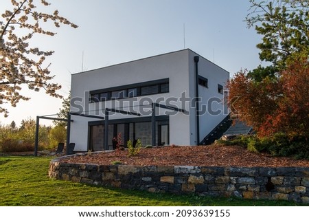 Modern, white, cube, elegant, minimalist style passive house with large panoramic windows, grey shutters in maintained garden in sunset warm light. Wooden terrace. Garden furniture.
