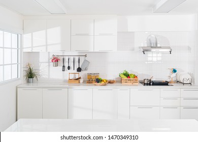 Modern White Kitchen With Counter And White Details, Minimalist Interior With Sunlight In Daytime. Full Set Of Kitchen Equipment, Pan, Pot, Electric Hob, Flipper, Vegetable, Fruit.