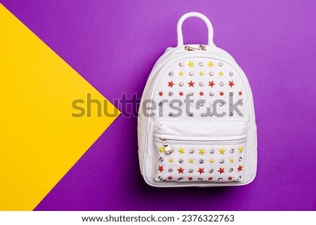 Modern white leather women's backpack with a front pocket covered with white, red, and yellow rivets-stars and rhinestones on a purple-yellow graphic background. Design for a fashion blog. Retail.