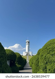 A modern white observation tower stands proudly at the end of a path lined with perfectly manicured green bushes. The sky is clear blue with a single fluffy cloud floating nearby, adding a dreamy touc