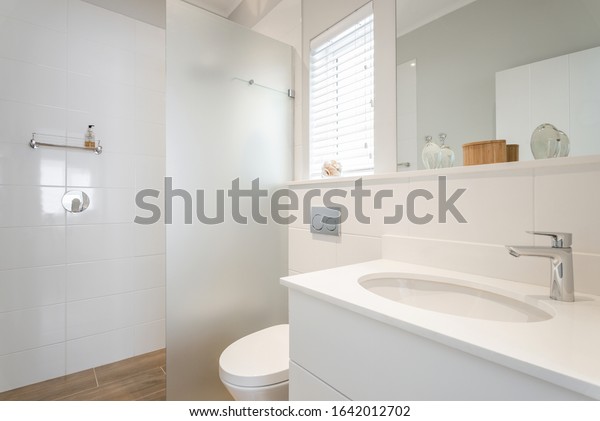 Modern white\
tiled bathroom with glass divided shower, hand basin and toilet,\
with decorative items and\
window
