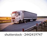 A modern white truck with a tilt semi-trailer transports cargo against the backdrop of a field and sunset in the summer. Transportation of building materials