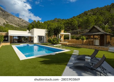 Modern white villa with a rectangular pool, sun loungers, stepping stones across a manicured lawn, and a dramatic mountain backdrop