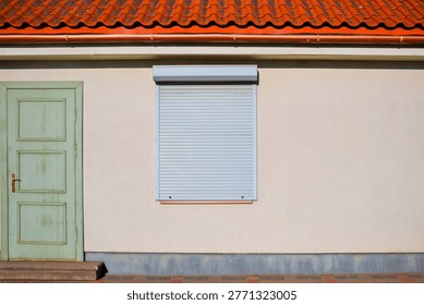 Modern window roller shutter on a house wall with red tile roof and green wooden door. Security and protection concept for residential building. Exterior house facade with copy space.