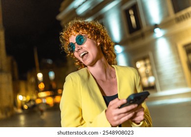 Modern woman in colorful casual clothes and blue sunglasses using smartphone laughing in the city at night - Powered by Shutterstock