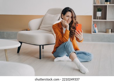 Modern Woman, Holding Smartphone, Taking Selfie Photo Or Chatting By Video Call, Sitting On Heated Floor In Living Room. Female Wearing Earphones Communicates Online Using Mobile Phone App At Home.