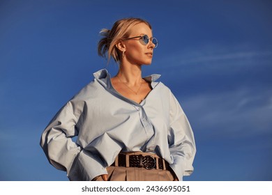 Modern woman in an oversized shirt and sunglasses stands outdoors against the sky. Chic, contemporary, and stylish, capturing of summer freedom.