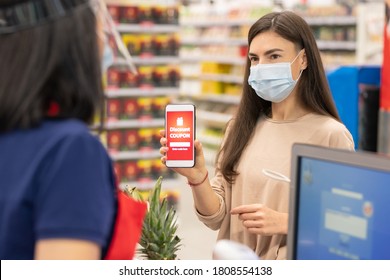 Modern Woman Wearing Mask On Face Using Digital Discount Coupon On Her Smartphone In Supermarket