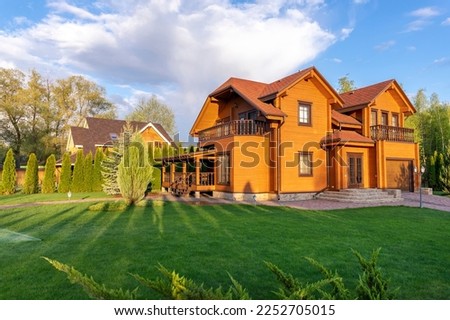 Modern wooden eco house villa facade luxury big house. Timber cottage with with green lawn water sprinkler, paved footpath and blue sky background. Landscaping design, garden watering and maintenance