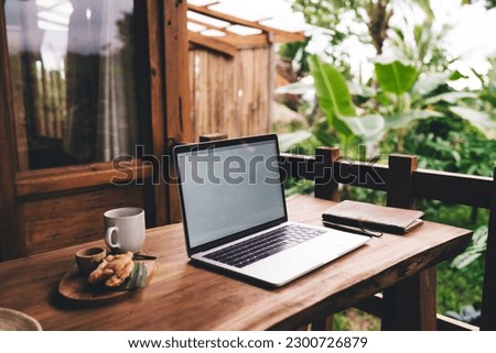 Modern wooden table placed with laptop with blank screen in daylight illuminating keypad panel and cup of coffee croissant served on tray while notepad and pen available at side