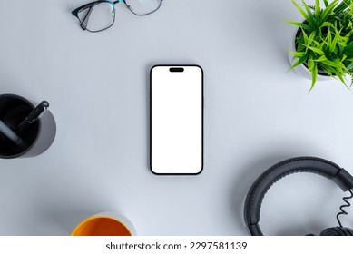 Modern work environment, with a blank phone screen islated for mockup, set among office essentials like a headset, plant, glasses, pens, and a coffee mug. Top view, flat lay composition - Powered by Shutterstock