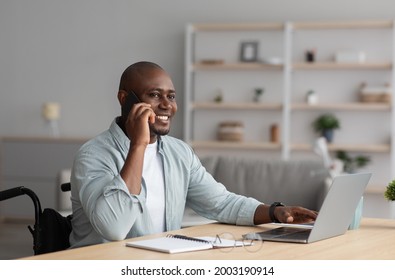 Modern Work From Home, Gadgets For Communication With Client, New Normal, Be Safe. Happy Adult Guy Disabled In Wheelchair Speaks By Phone At Workplace With Laptop In Living Room Interior, Empty Space