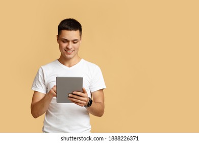 Modern work and leisure time with mobile gadget. Attractive friendly european young guy blogger using digital tablet, surfing in internet, isolated on beige background, studio shot, free space - Powered by Shutterstock