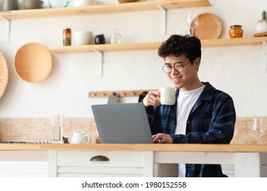 Modern Worker In Self-isolation, Home Office And Work During Covid-19 Quarantine. Smiling Young Asian Male With Cup, Drinking Coffee And Working On Laptop At Table In Kitchen Interior, Free Space