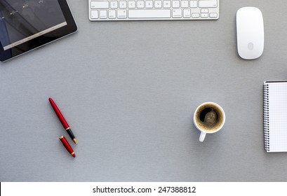 Modern Working Place On Grey Wood Table. From Above View On Well Equipped Working Place With Tablet PC, Computer Mouse And Keyboard, Red Pen, Cup Of Fresh Coffee, And Blank Notepad