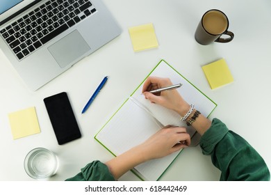 Modern Workplace Flat Lay. Top View Of Home Office Desk With Laptop, Smartphone, Cup Of Coffee And Water And Female Hands Writing In Note Pad